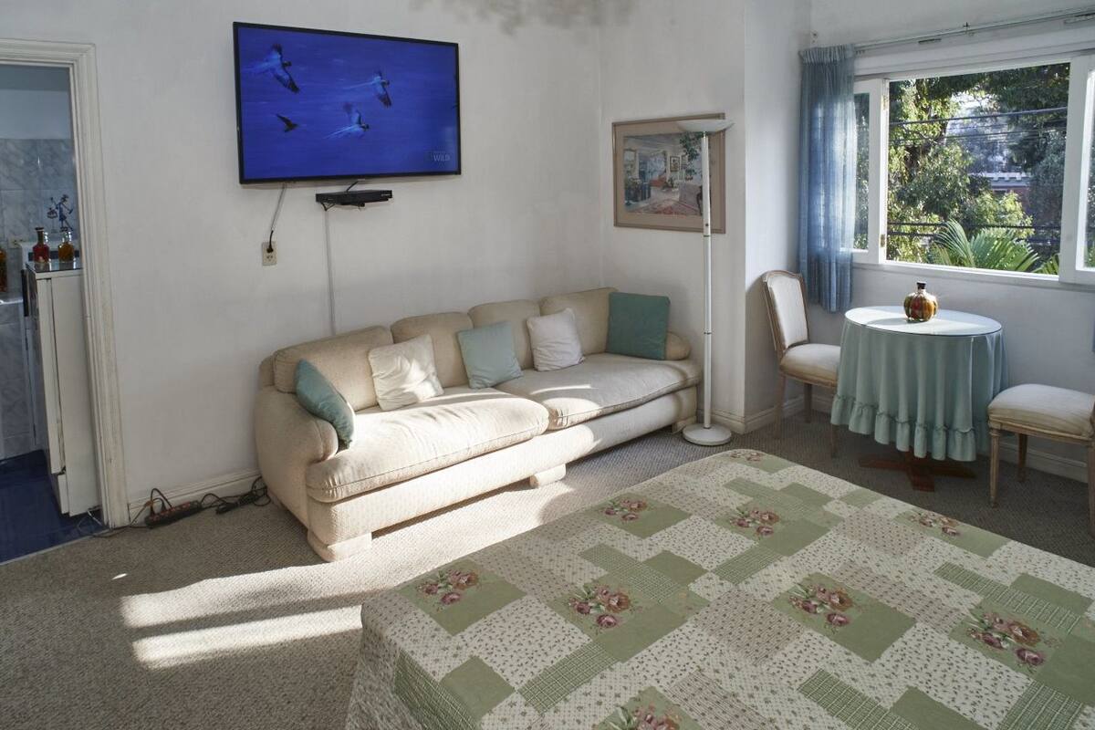 Ein Wohnzimmer mit einem beigefarbenen Sofa, einem runden Tisch mit blauer Tischdecke, einem Fernseher an der Wand und einem Fenster mit Blick auf Grünflächen.