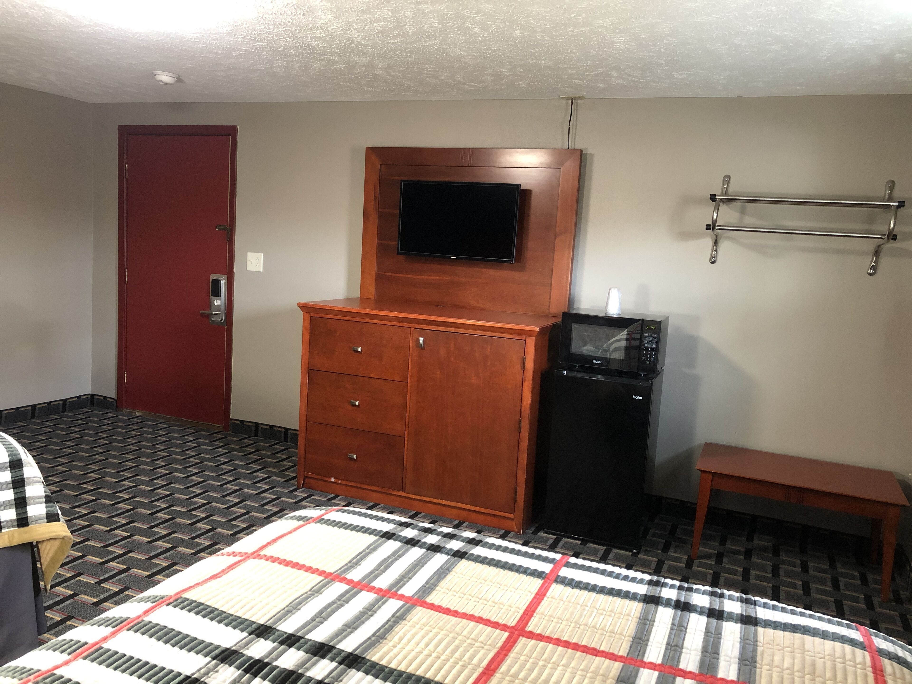 A hotel room with a wooden dresser, a black refrigerator, a television mounted above the dresser, a red door, and a patterned rug.