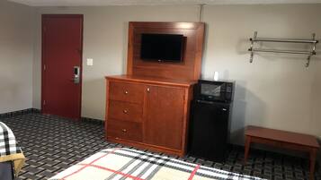 A hotel room with a wooden dresser, a black refrigerator, a television mounted above the dresser, a red door, and a patterned rug.