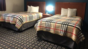 A hotel room with two beds, each with a plaid bedspread and white pillows. There is a nightstand with a lamp between the beds. The room has a patterned carpet and blue walls. - Royal Motel (Martinsville)