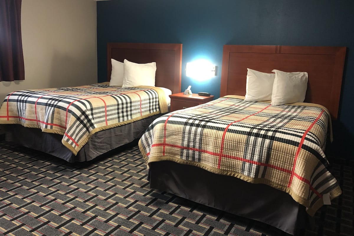 Hotellihuoneessa on kaksi sänkyä, joissa on molemmissa ruutukuviollinen päiväpeite ja valkoiset tyynyt. Sänkyjen välissä on yöpöytä, jossa on lamppu. Huoneessa on kuvioitu matto ja siniset seinät.