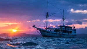 Una barca con più alberi e una cabina, che naviga sull'acqua al tramonto.