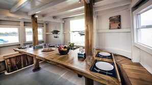 Zona pranzo con tavolo in legno apparecchiato per due persone, una ciotola di frutta e vista sull'acqua.