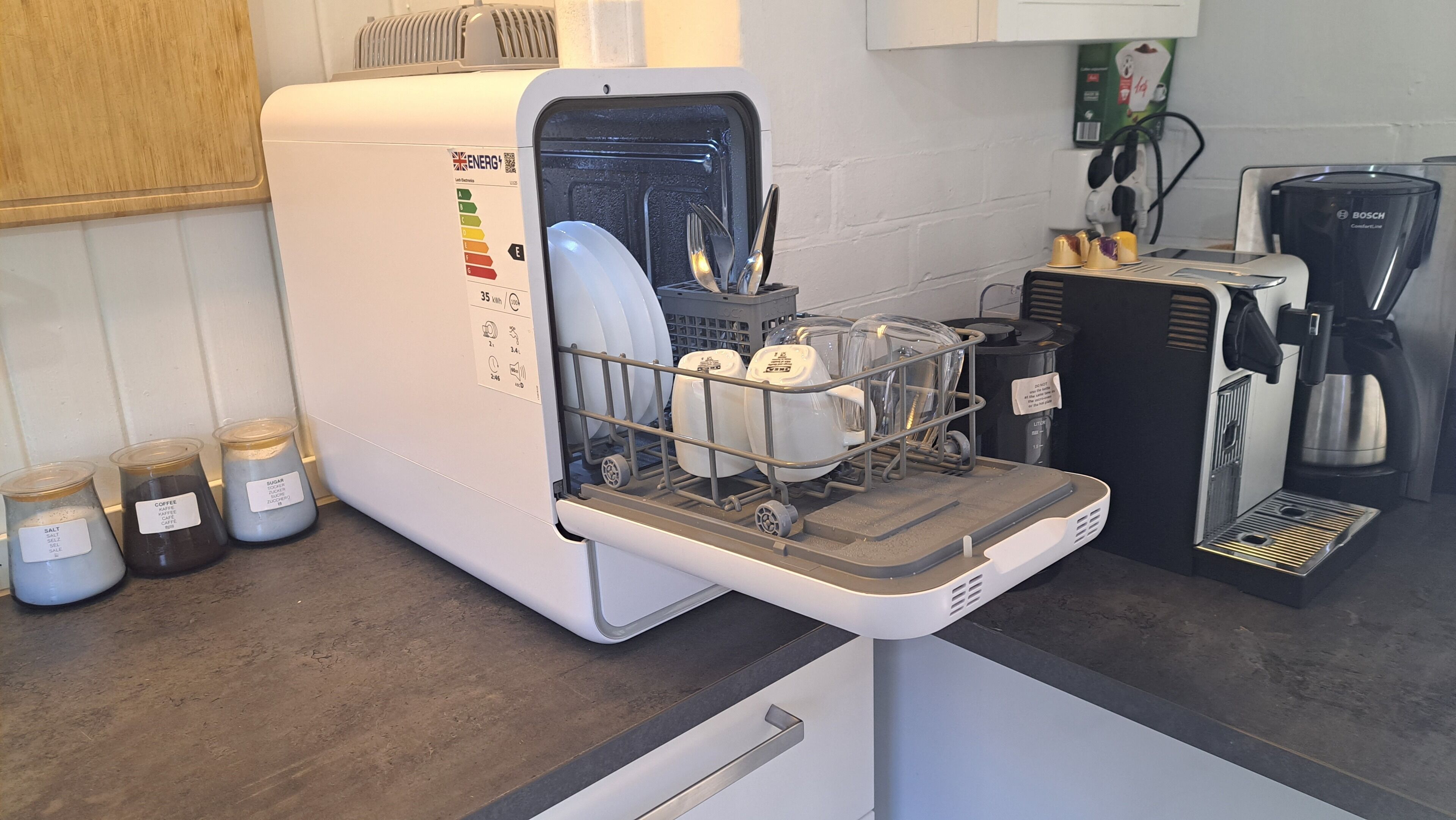 A compact kitchen with a built-in dishwasher, a coffee maker, and storage jars labeled with contents like salt and coffee.