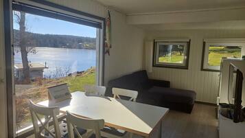 A room with a large window overlooking a lake, a white table with chairs, a dark sofa, and a small TV on a stand.