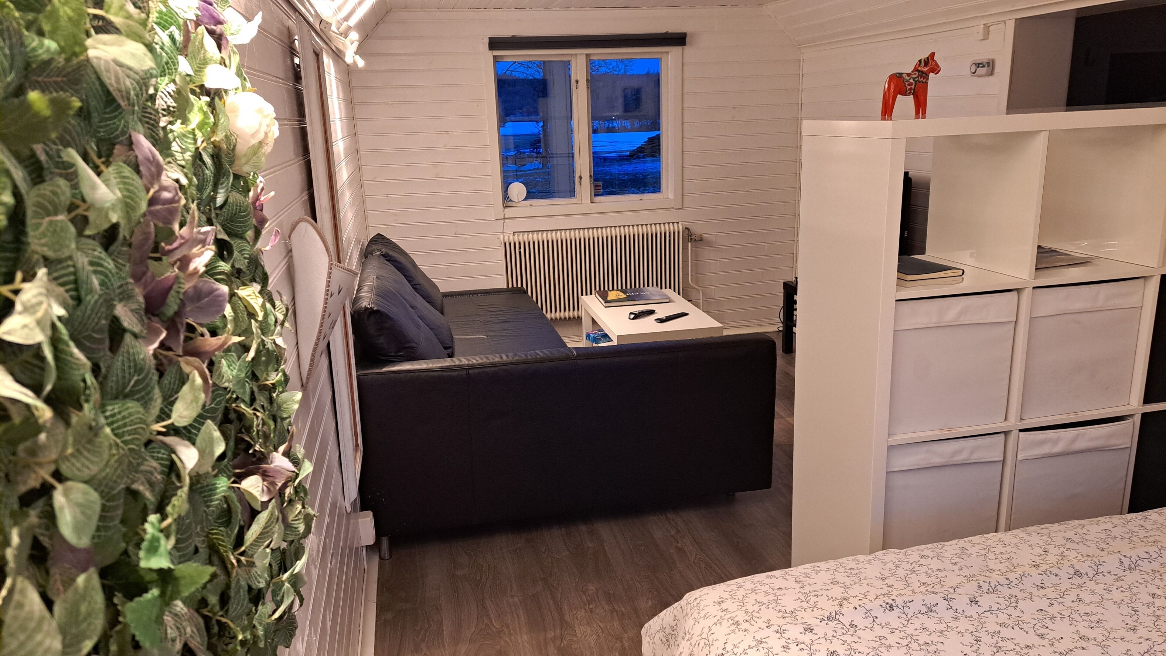 A compact room with a bed, a small table with a book and remote, a radiator, and a window with a view of the outside.