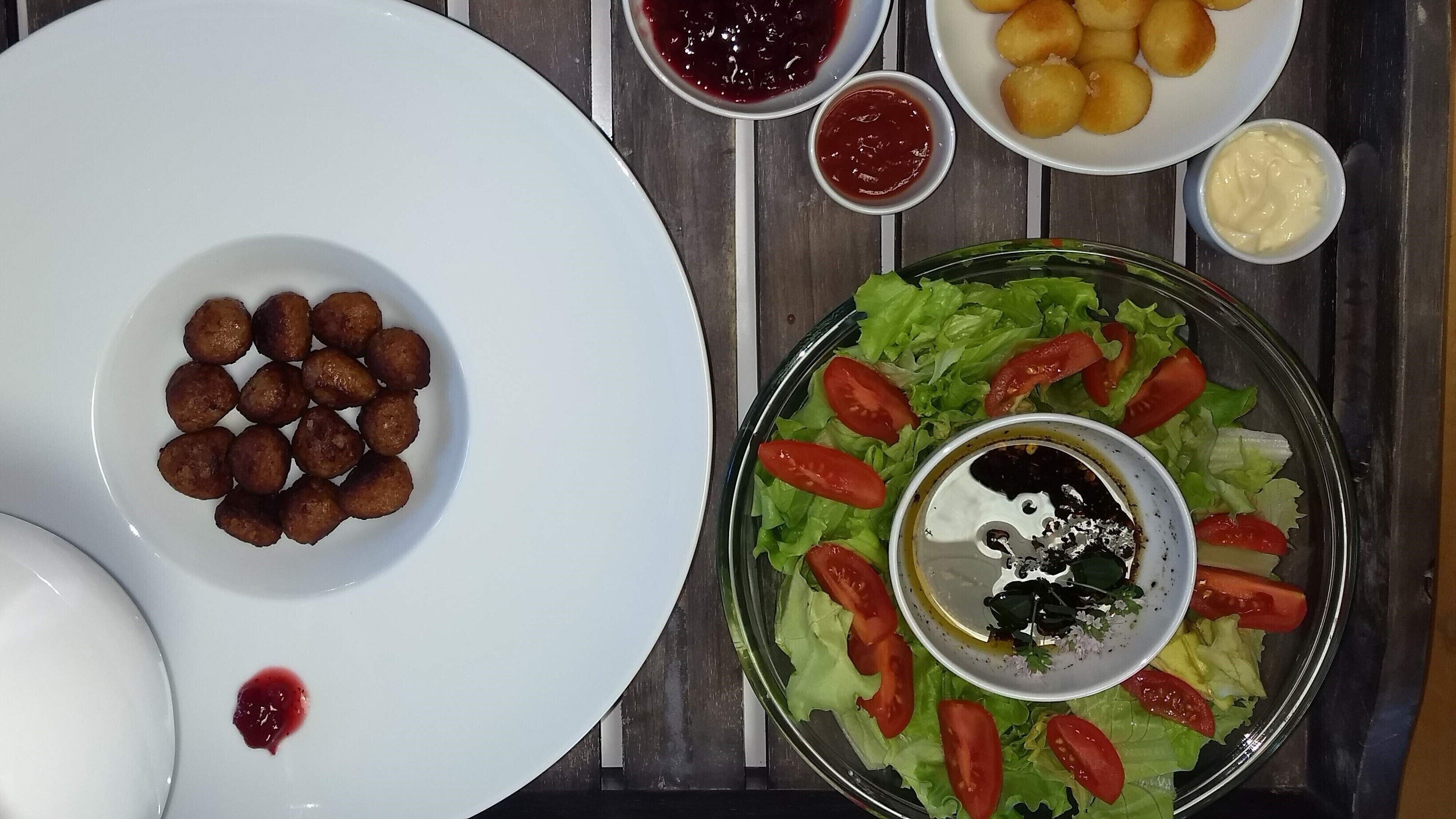 A table with a plate of round, golden-brown fried items, a salad with lettuce and tomatoes, and two small bowls of red and white sauces.