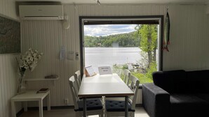 Una habitación con una mesa de comedor y sillas, una mesita blanca con un jarrón de flores, un sofá negro y vistas a un lago a través de una puerta.