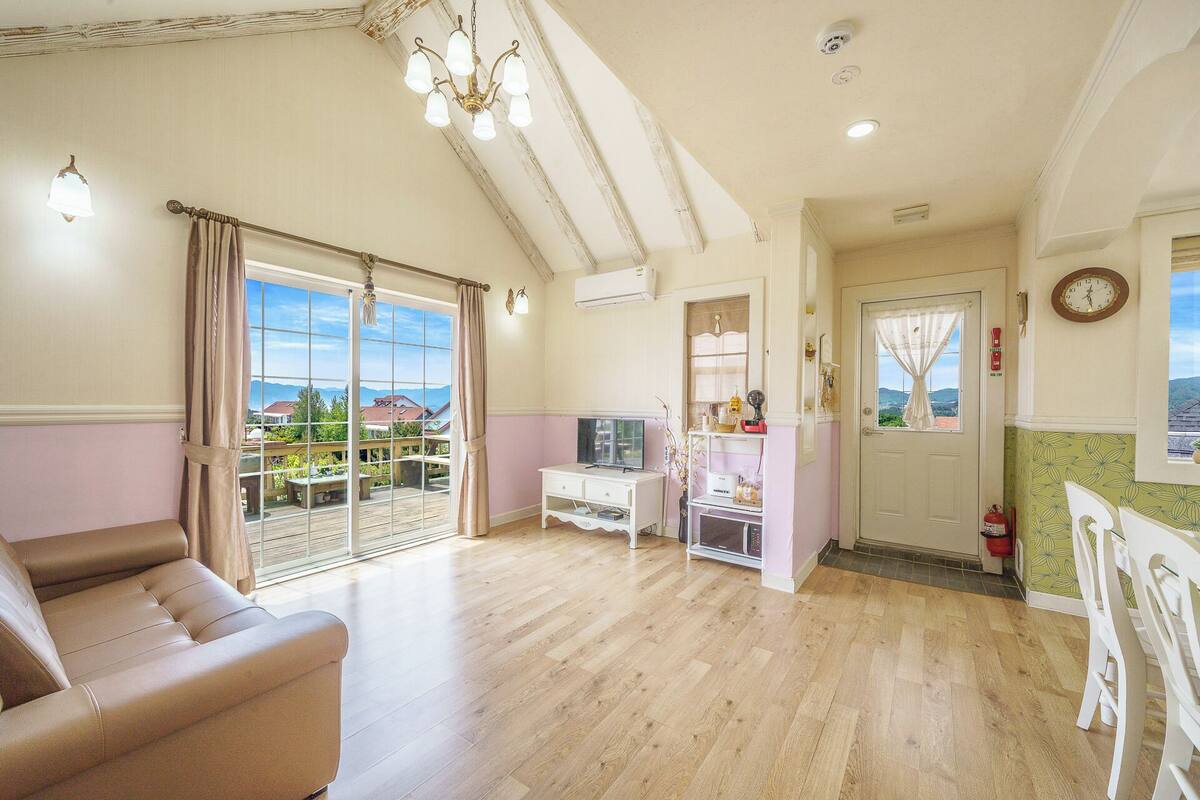 Amplia sala de estar con un gran ventanal corredero que da a un balcón, un mueble blanco para televisor y una puerta con vistas al exterior.