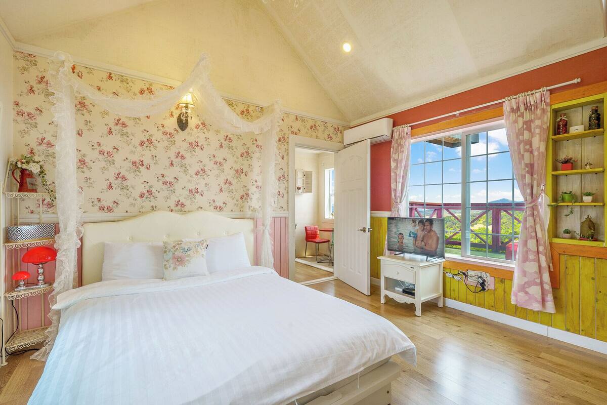 Un dormitorio con una cama grande, una mesita de noche, una lámpara, un ventanal con vistas y un estante con objetos decorativos.
