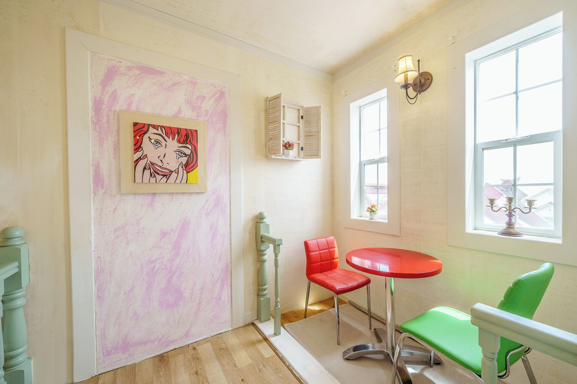A room with a pink wall, a framed artwork, a red chair, a green chair, a red table, and a window with a view.