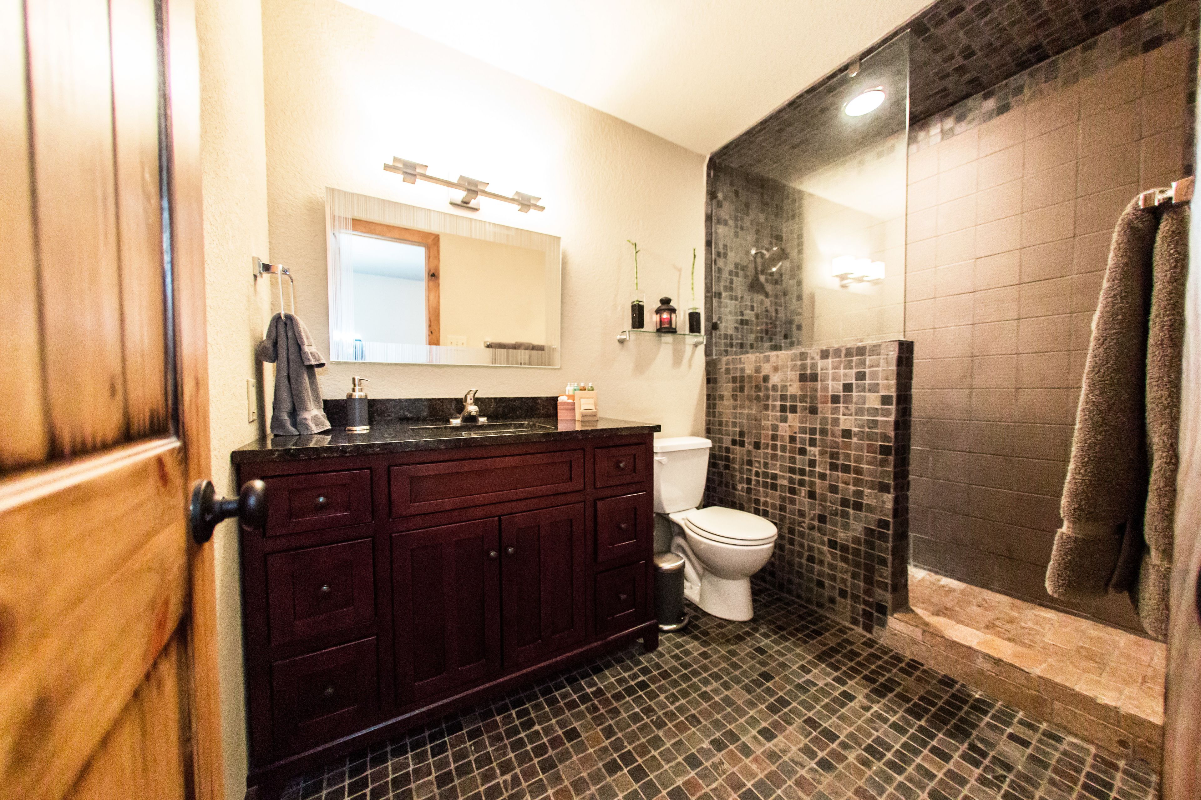 A bathroom with a dark tiled shower, a white toilet, and a wooden vanity with a mirror above it.