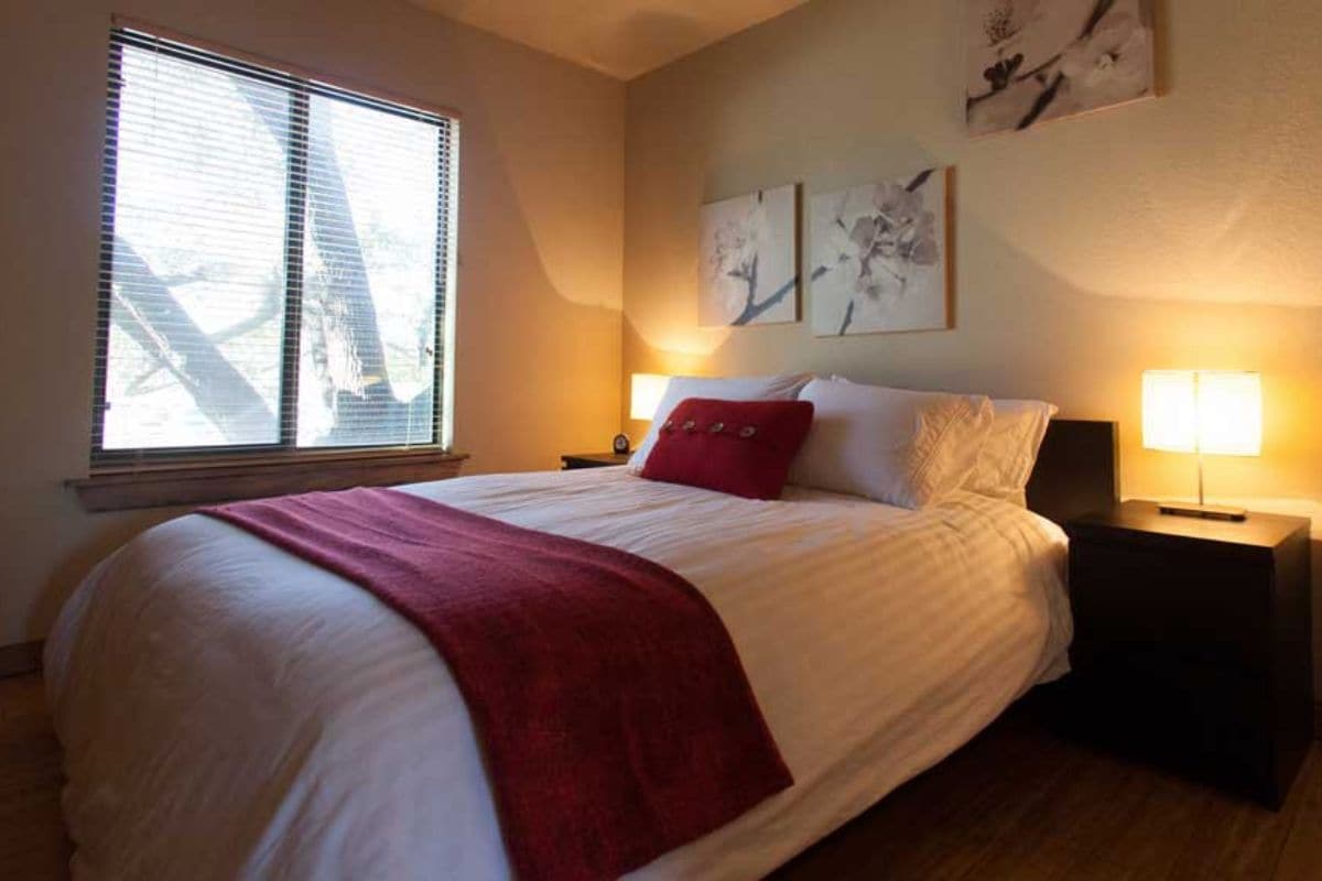 A neatly made bed with a red throw blanket, a nightstand with a lamp, and artwork on the wall.