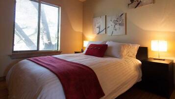 A neatly made bed with a red throw blanket, a nightstand with a lamp, and artwork on the wall.