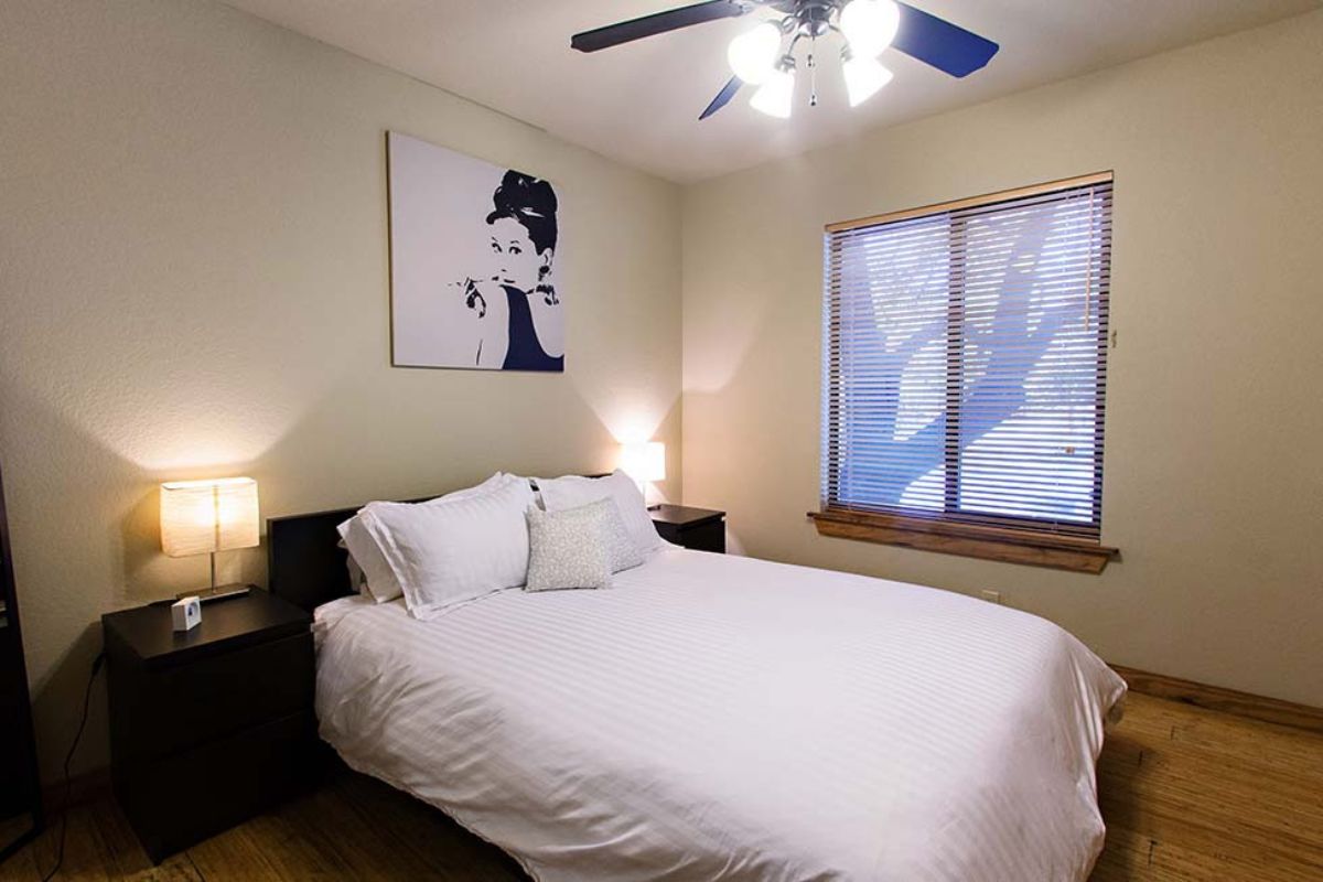 A bedroom with a bed, nightstands, a ceiling fan, a wall-mounted artwork, and a window with blinds.