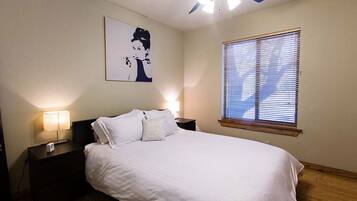 A bedroom with a bed, nightstands, a ceiling fan, a wall-mounted artwork, and a window with blinds.