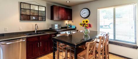 A modern kitchen with dark wood cabinets, stainless steel appliances, a black granite countertop, and a dining area with wooden chairs.