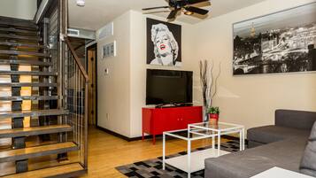 A modern living room with a staircase, a red entertainment unit, a flat-screen TV, a gray sofa, and a cityscape painting.
