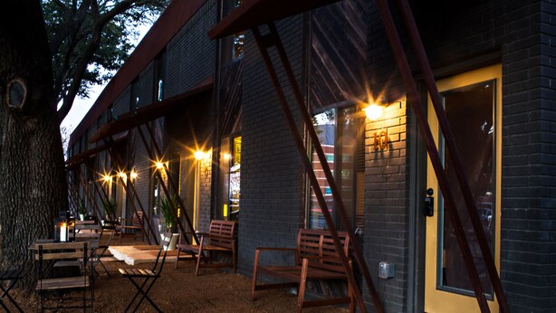 An outdoor seating area with wooden chairs and tables, a brick building with a yellow door, and a tree in the background. - 1700 South 2nd (Waco)