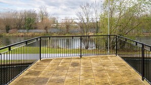 A paved deck with a black metal railing overlooking a body of water, surrounded by trees and a distant bridge. - Luxury 2 Bed Riverside (Bedford)