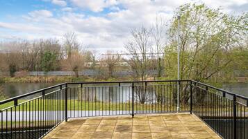 A paved deck with a black metal railing overlooking a body of water, surrounded by trees and a distant bridge.