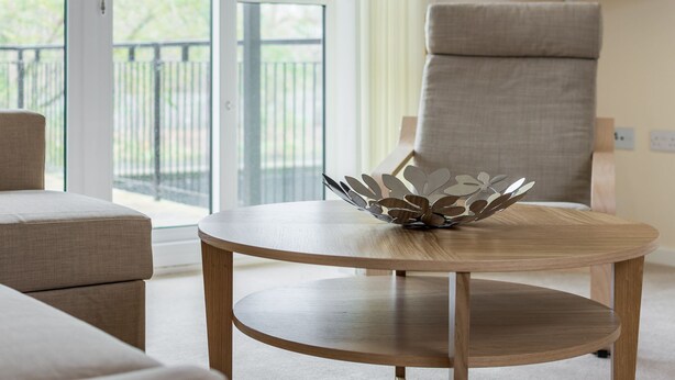 A modern living room with a round wooden table, a beige armchair, and a large window with a view of greenery. - Luxury 2 Bed Riverside (Bedford)