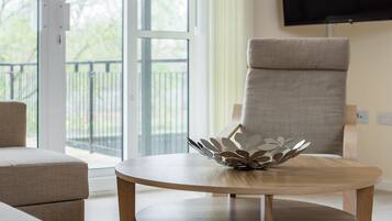 A modern living room with a round wooden table, a beige armchair, and a large window with a view of greenery.