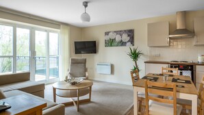 A modern living room with a sofa, a round coffee table, and a dining area with a wooden table and chairs. There is a TV mounted on the wall, a painting of flowers, and a balcony with a railing. - Luxury 2 Bed Riverside (Bedford)