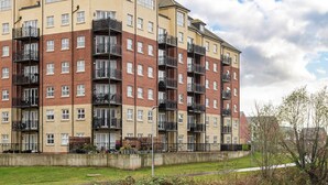 A multi-story residential building with balconies, a concrete retaining wall, and a grassy area in front. - Luxury 2 Bed Riverside (Bedford)