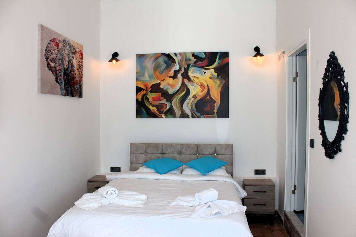 A neatly made bed with blue pillows, a large abstract painting above it, and a mirror on the wall.