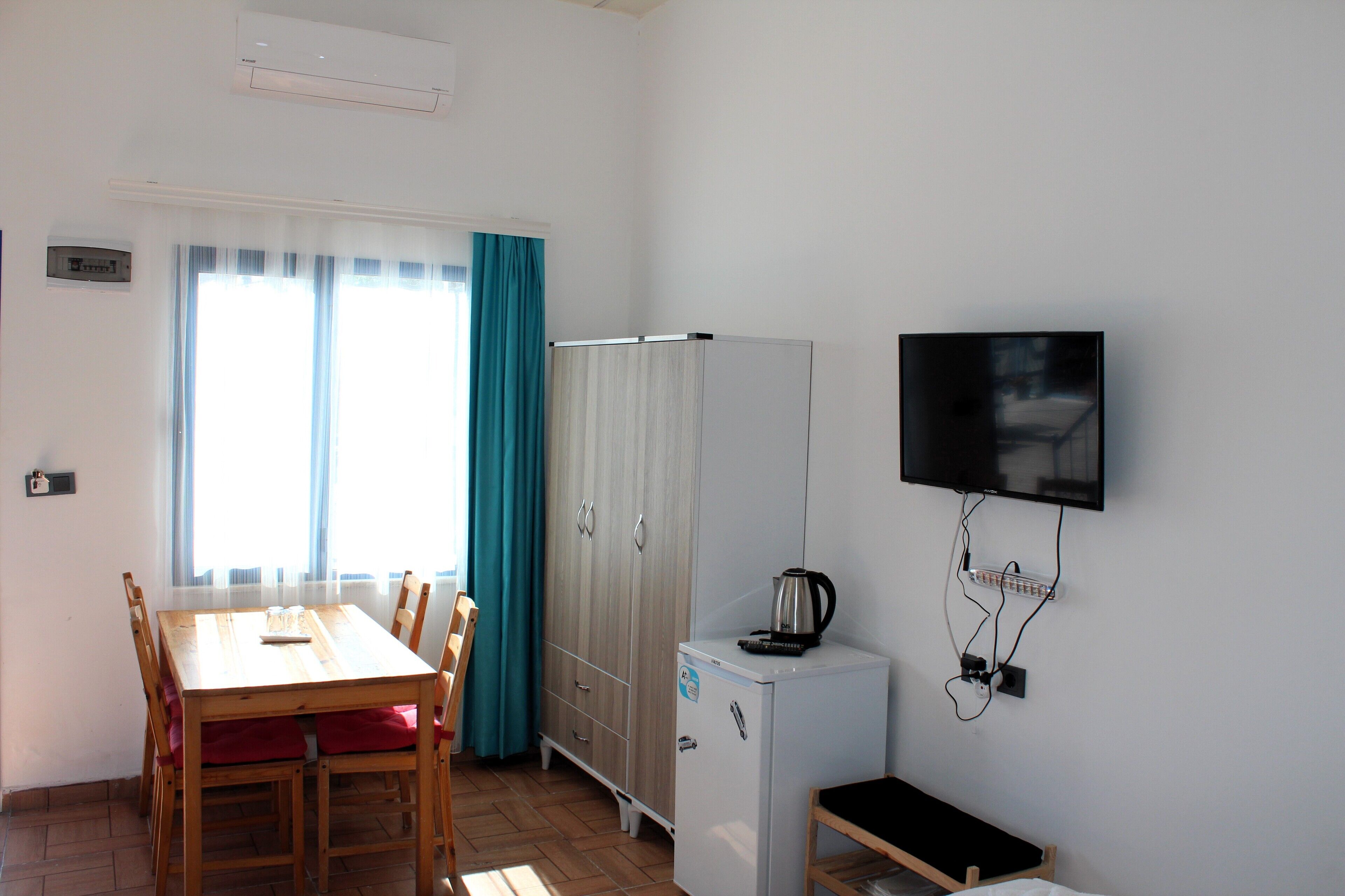 Una habitación pequeña con una mesa de comedor de madera, sillas, un armario blanco, un refrigerador blanco pequeño y un televisor montado.
