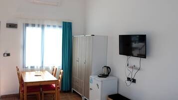 Una habitación pequeña con una mesa de comedor de madera, sillas, un armario blanco, un refrigerador blanco pequeño y un televisor montado.