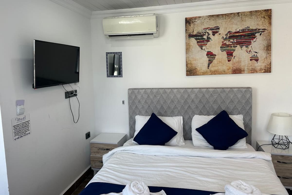A hotel room with a bed, a TV, an air conditioner, a world map artwork, and a bedside table with a lamp.