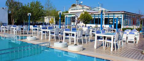 A poolside area with lounge chairs and tables, a clear blue pool, and a building with large windows and a balcony.