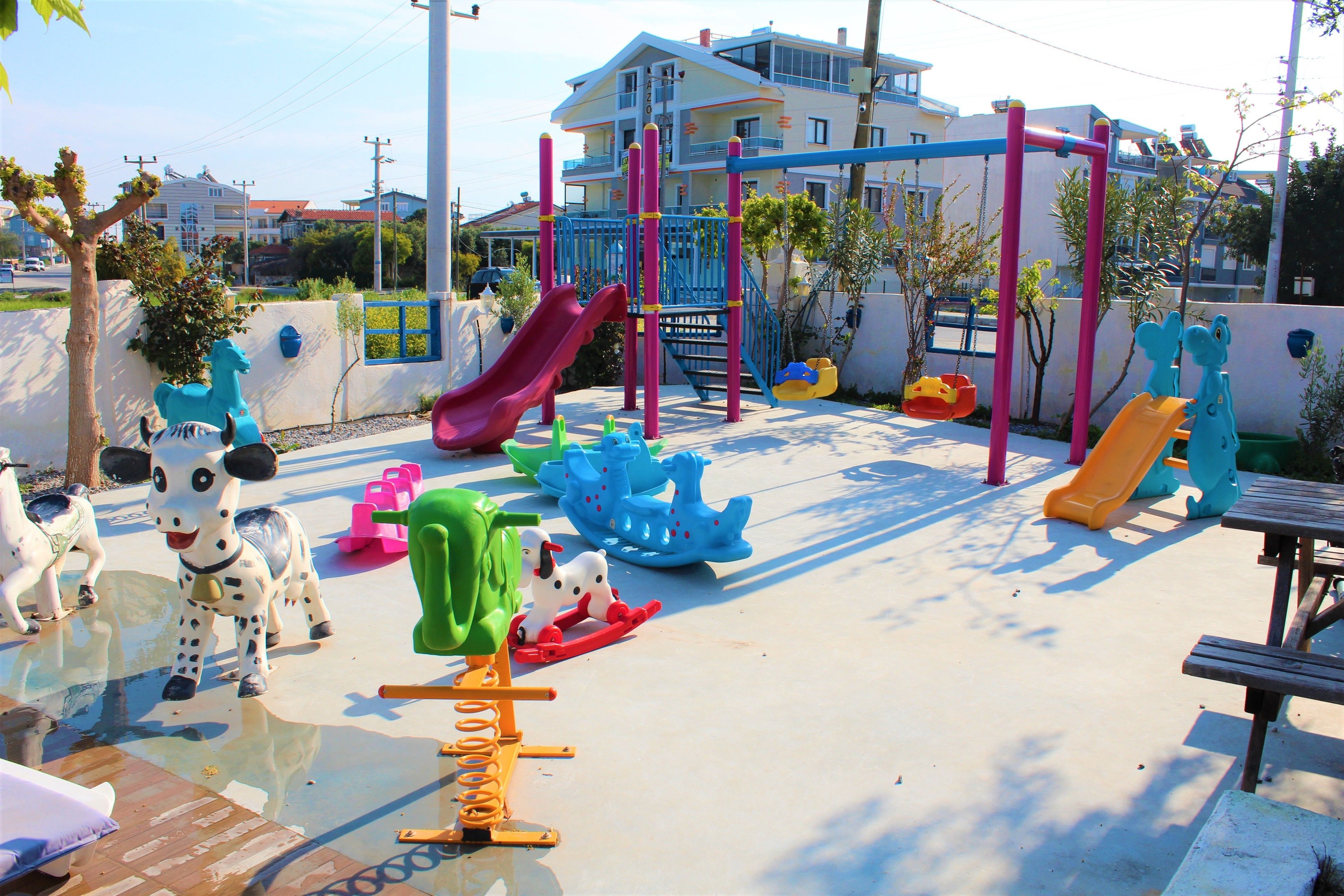 A playground with slides and climbing structures, surrounded by a white wall and trees.