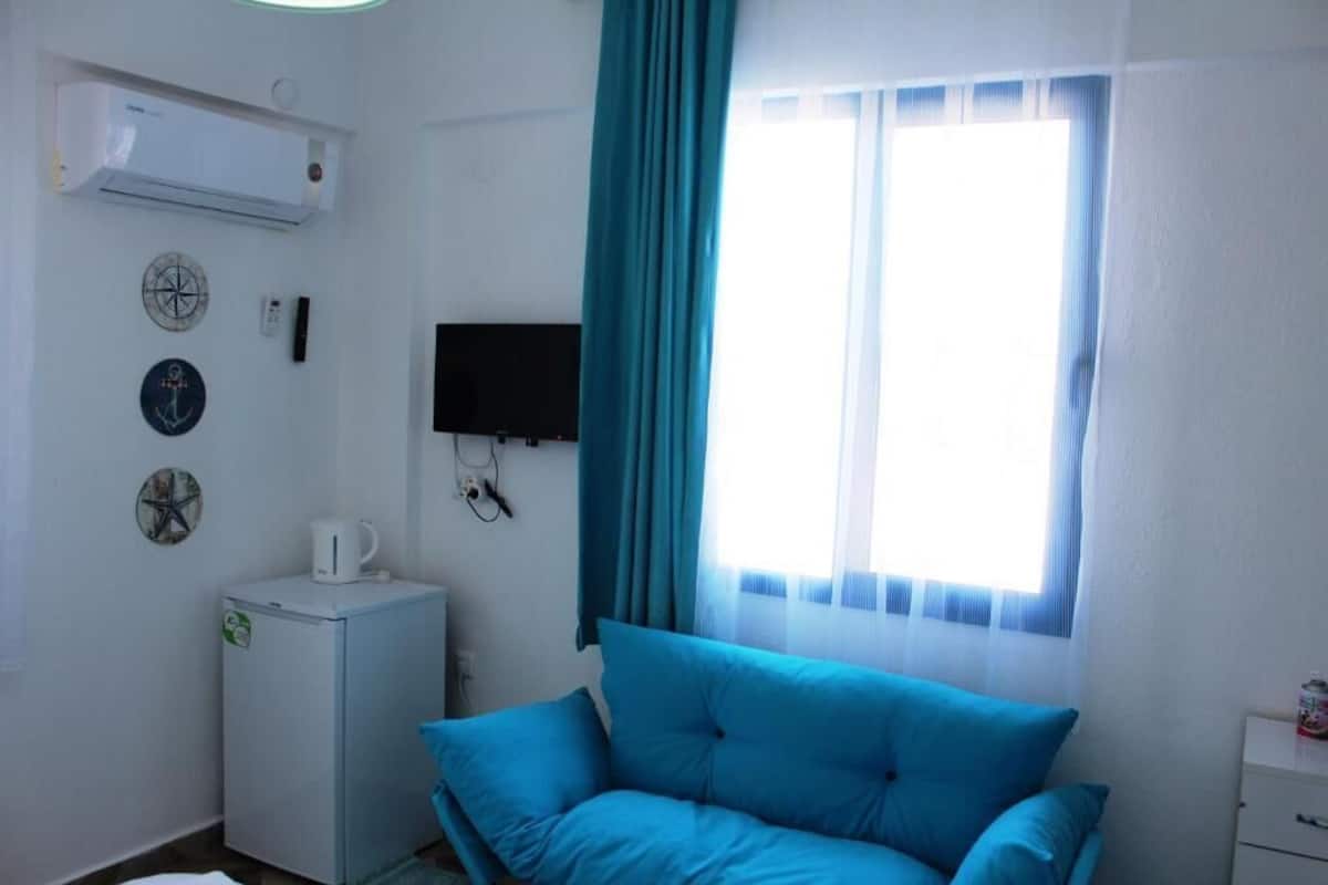 A small room with a blue sofa, a white mini-fridge, a television mounted on the wall, and a window with a blue curtain.