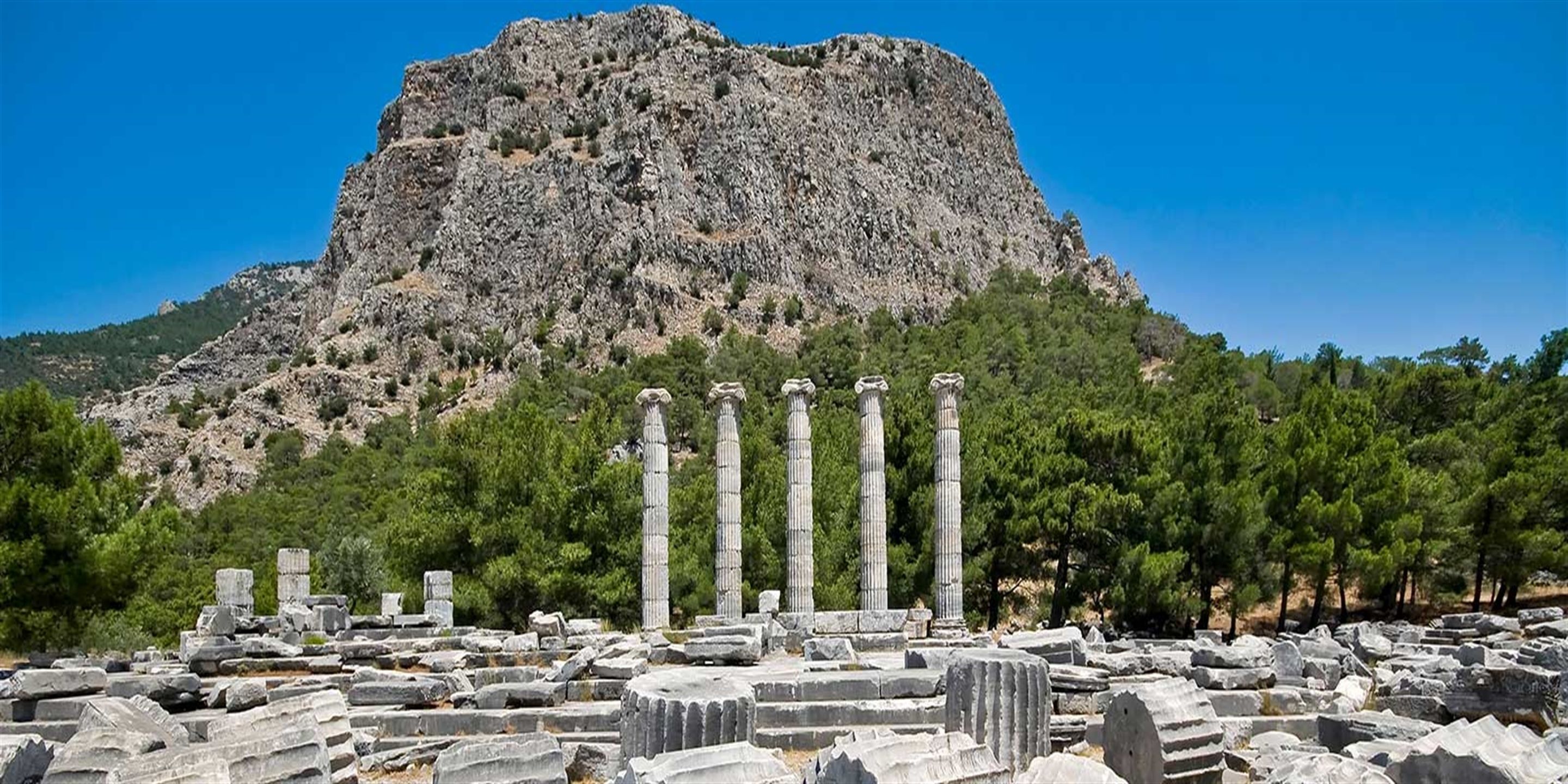 Ancient ruins with columns and stone blocks, set against a mountainous backdrop.