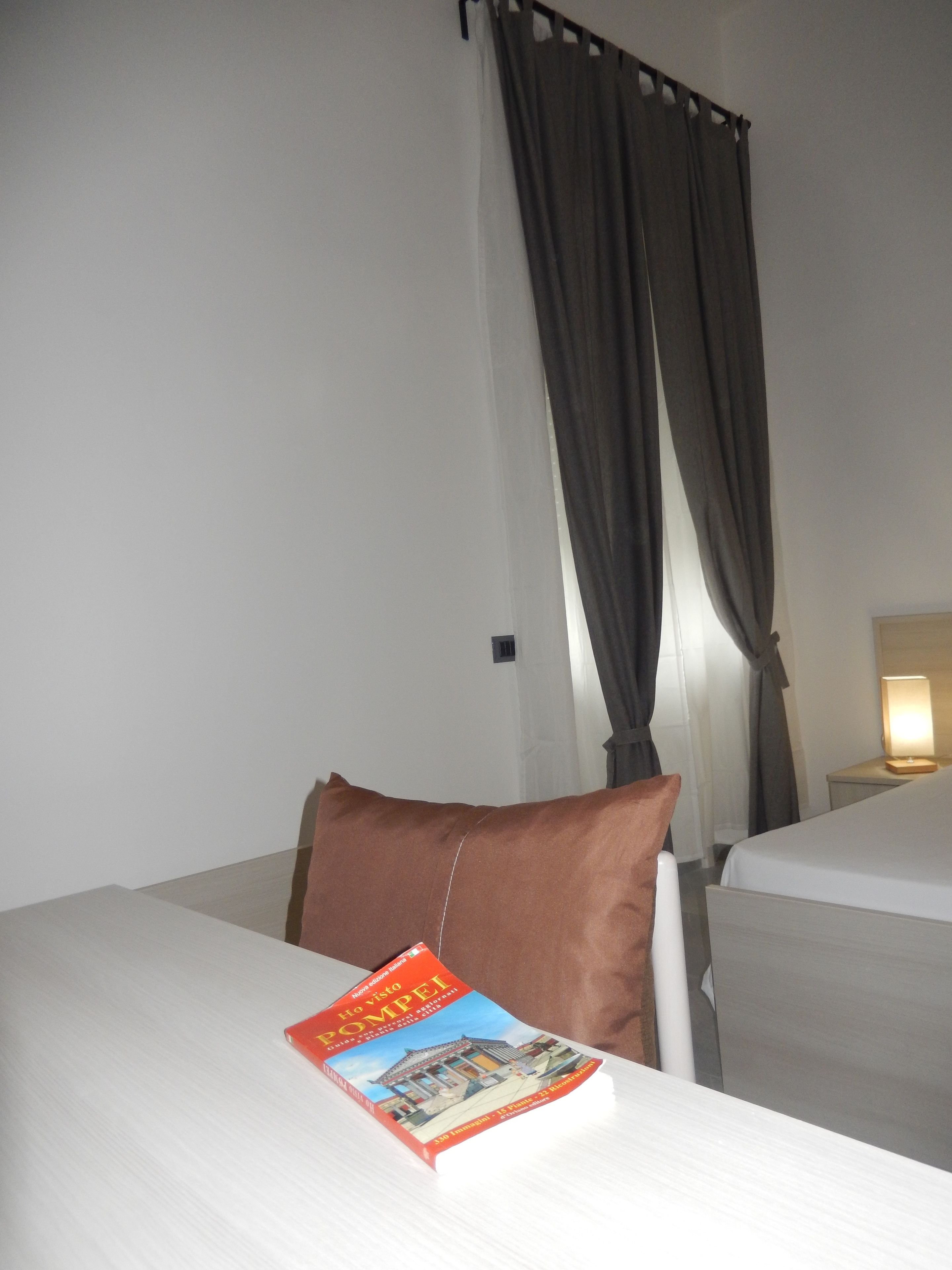 A hotel room with a bed, a pillow, a book, a lamp, and curtains.