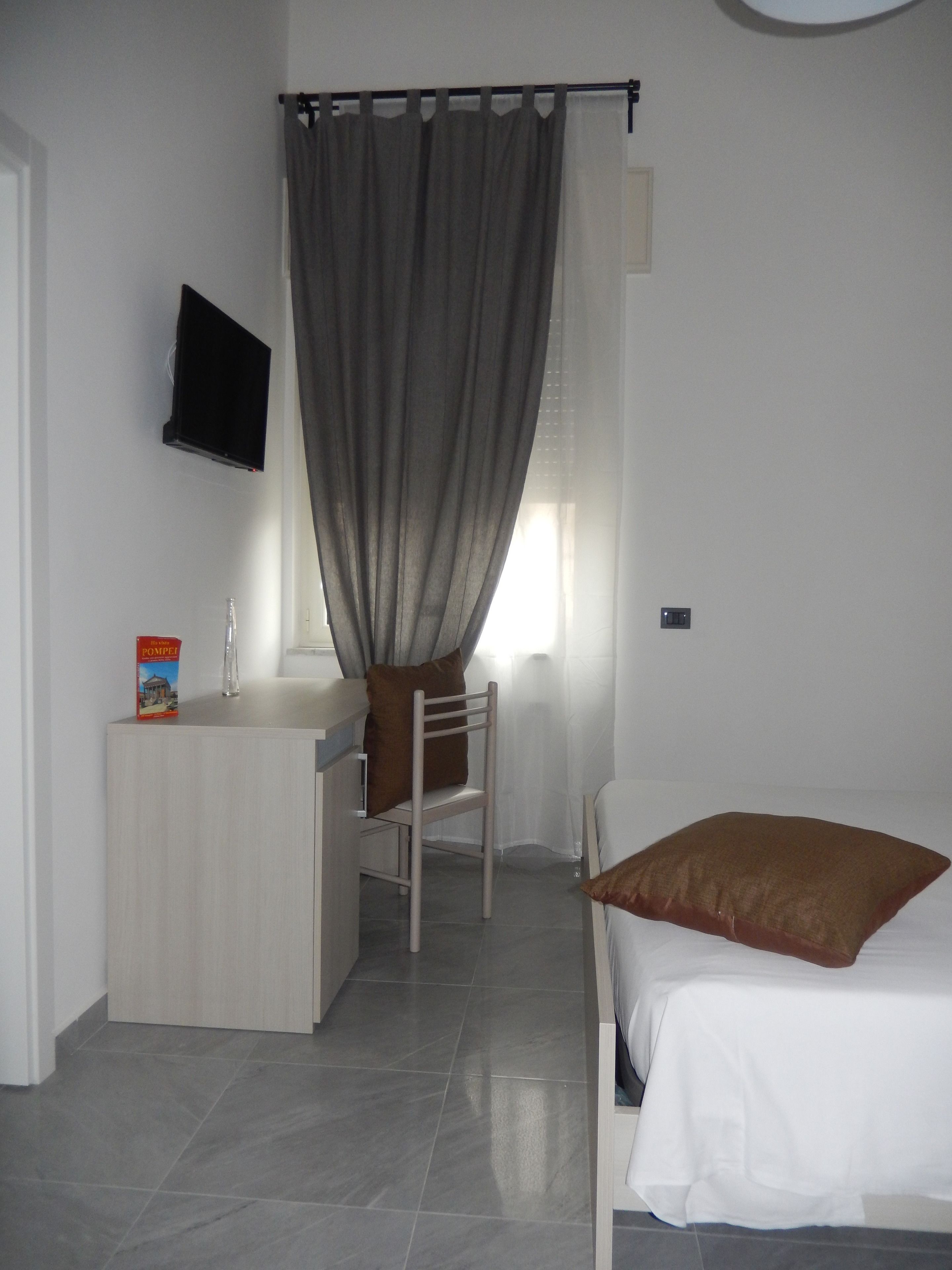 Ein Schlafzimmer mit Bett, Schreibtisch, Stuhl, einem an der Wand befestigten Fernseher und einem Fenster mit Vorhängen.