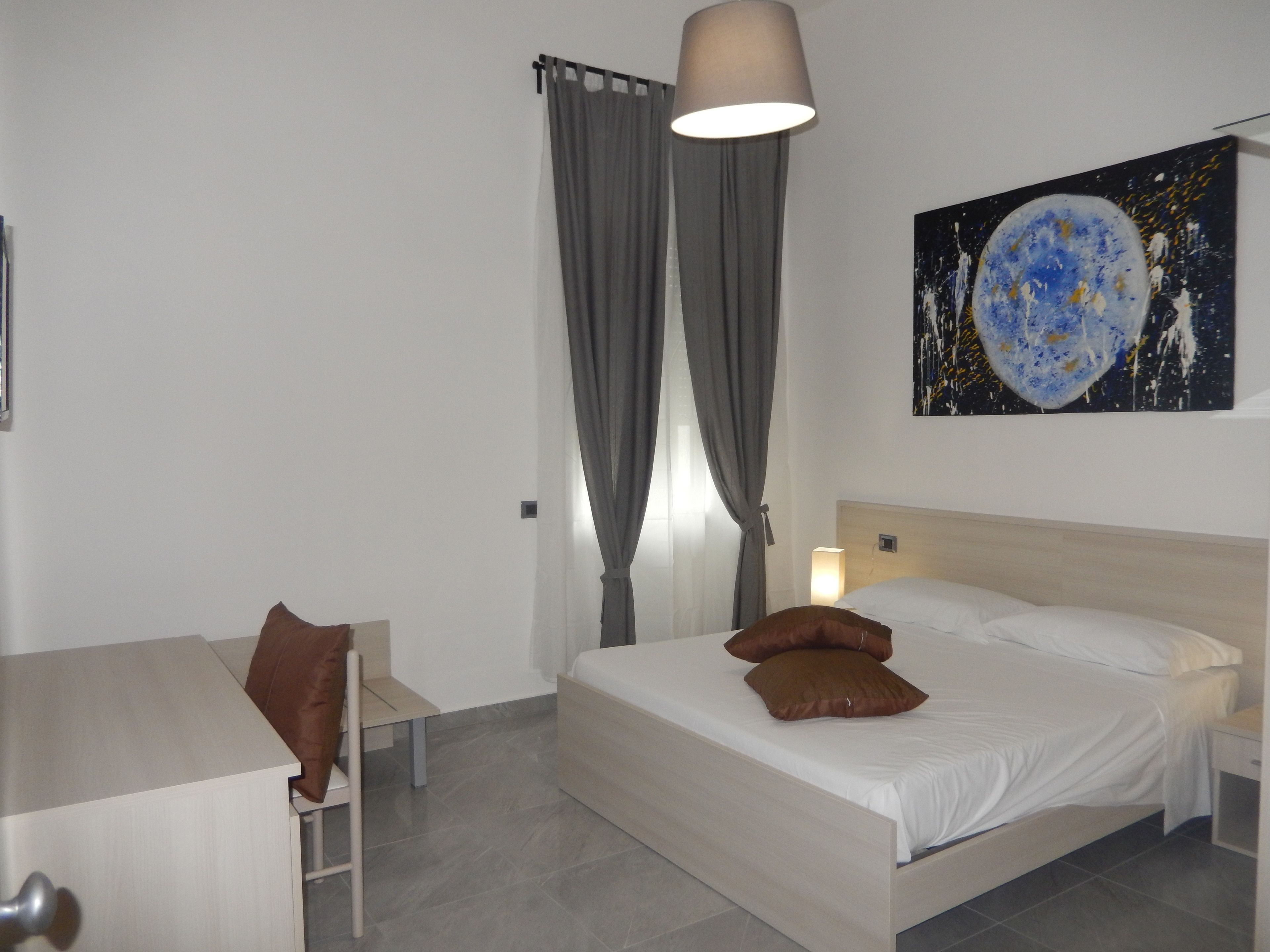 A bedroom with a bed, a desk, a chair, a lamp, and a painting on the wall.
