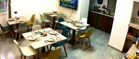 Un comedor con varias mesas y sillas, un televisor montado en la pared y una cocina visible a través de un arco.
