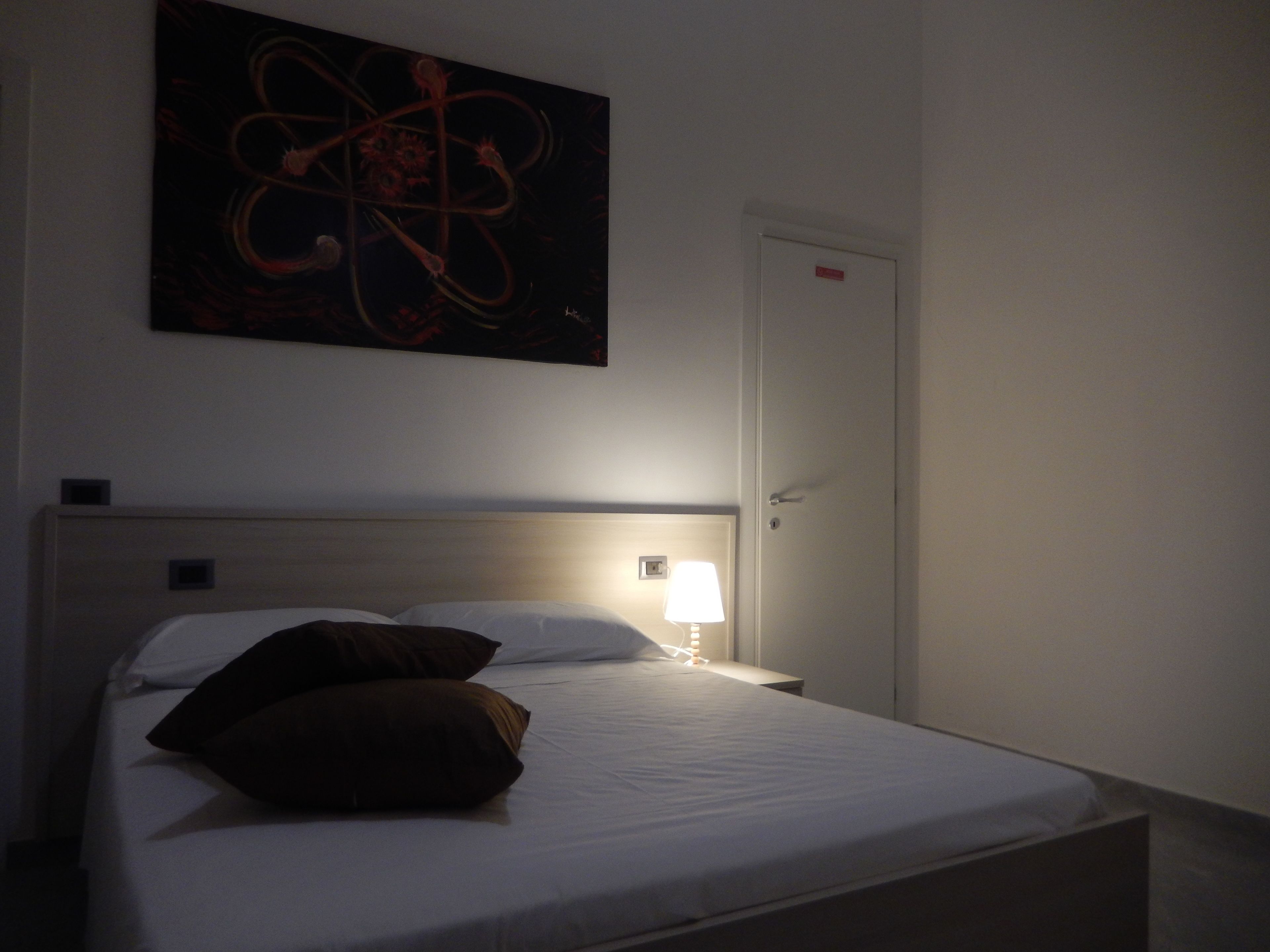A single bed with white bedding and two pillows, a bedside lamp, and a wall-mounted abstract painting.