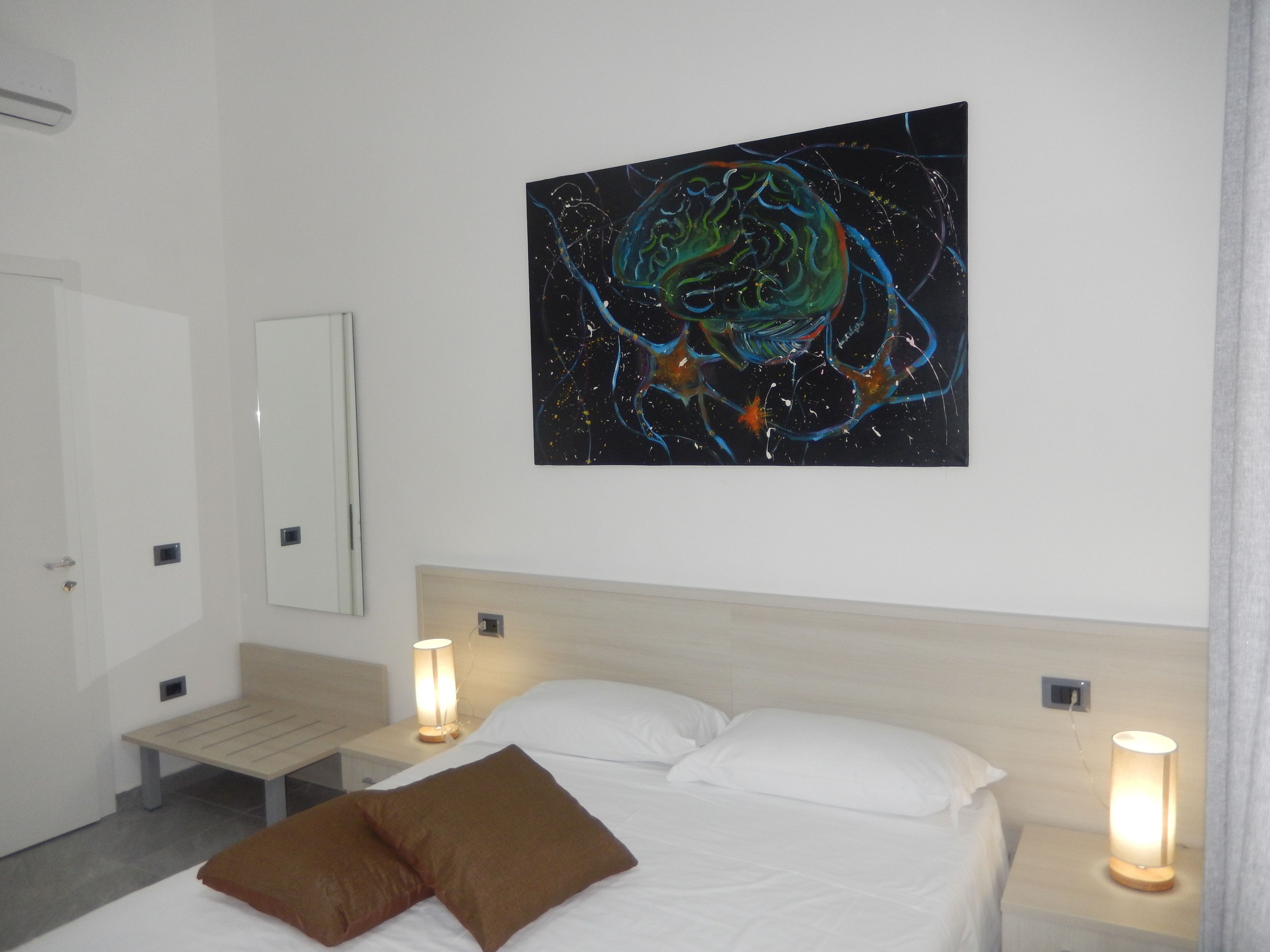 A hotel room with a bed, two bedside tables with lamps, a large abstract painting on the wall, and a mirror.
