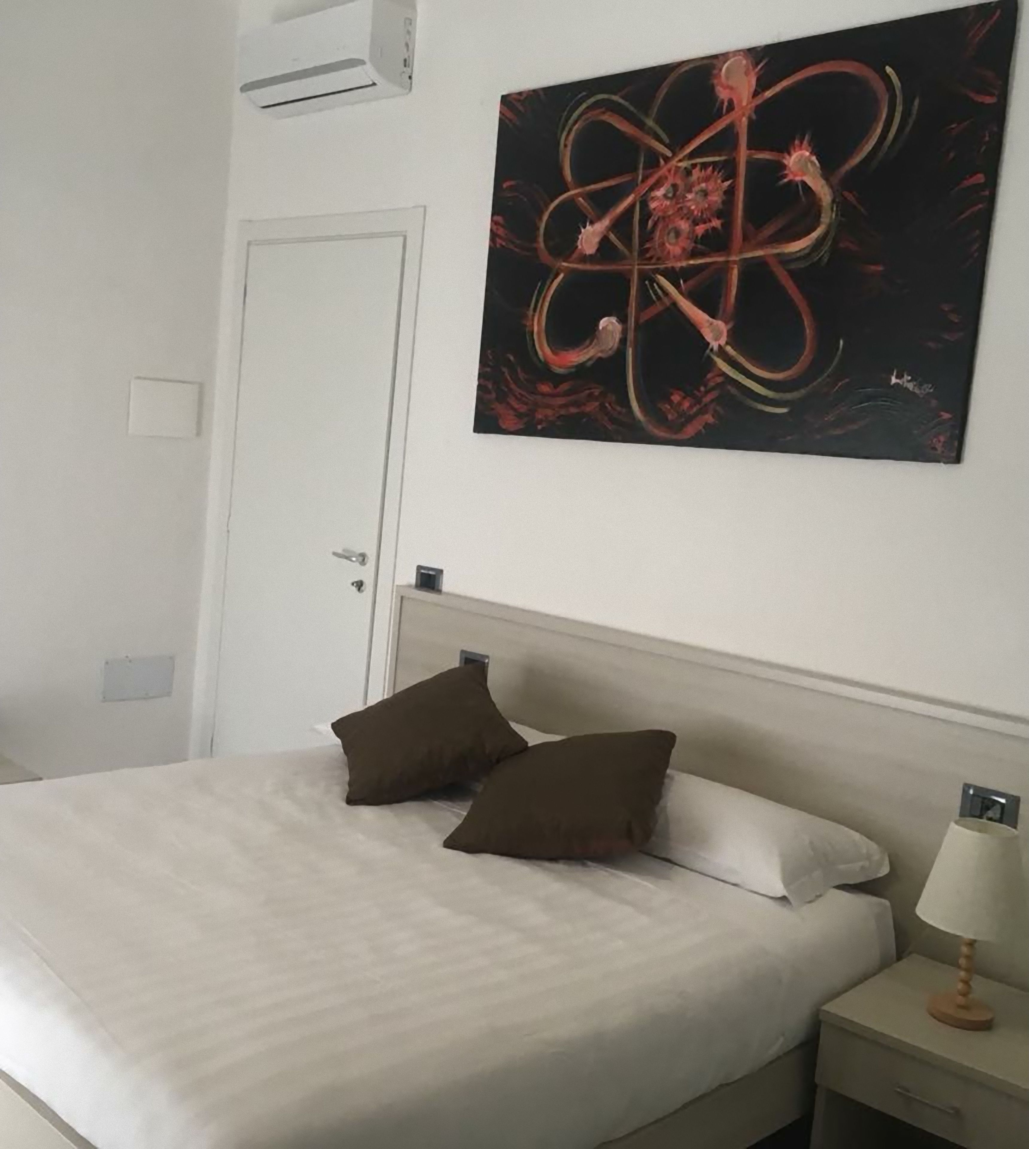 A bedroom with a bed, two pillows, a nightstand with a lamp, and a wall-mounted painting.