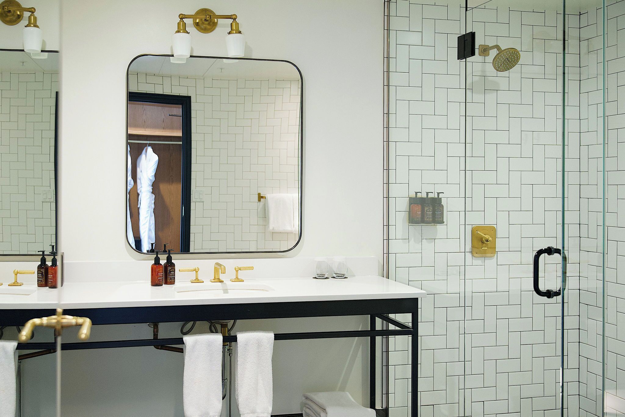 A bathroom with a white tiled wall, a large mirror, a sink with gold fixtures, and a glass shower enclosure.