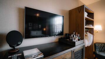 A hotel room with a large flat-screen TV mounted on the wall, a wooden cabinet with a mirror, a bedside table with a lamp, and a comfortable armchair.