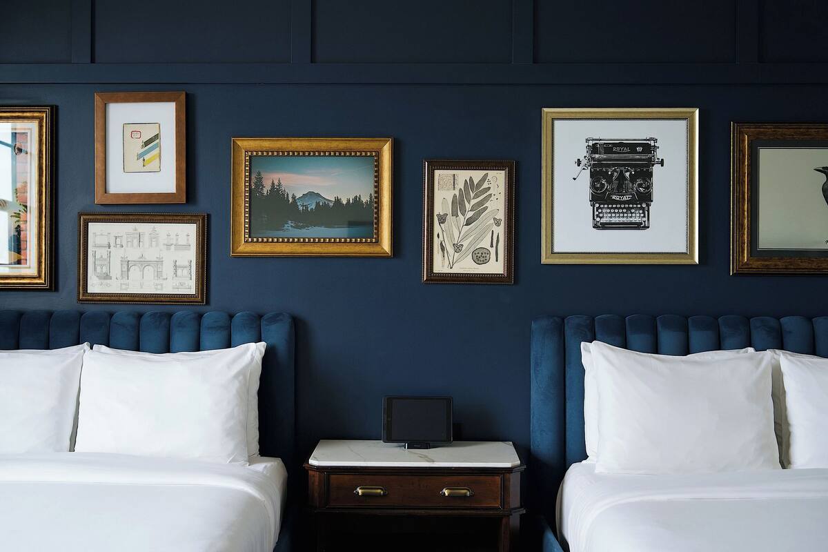A hotel room with two beds, a nightstand, and a wall decorated with framed pictures.