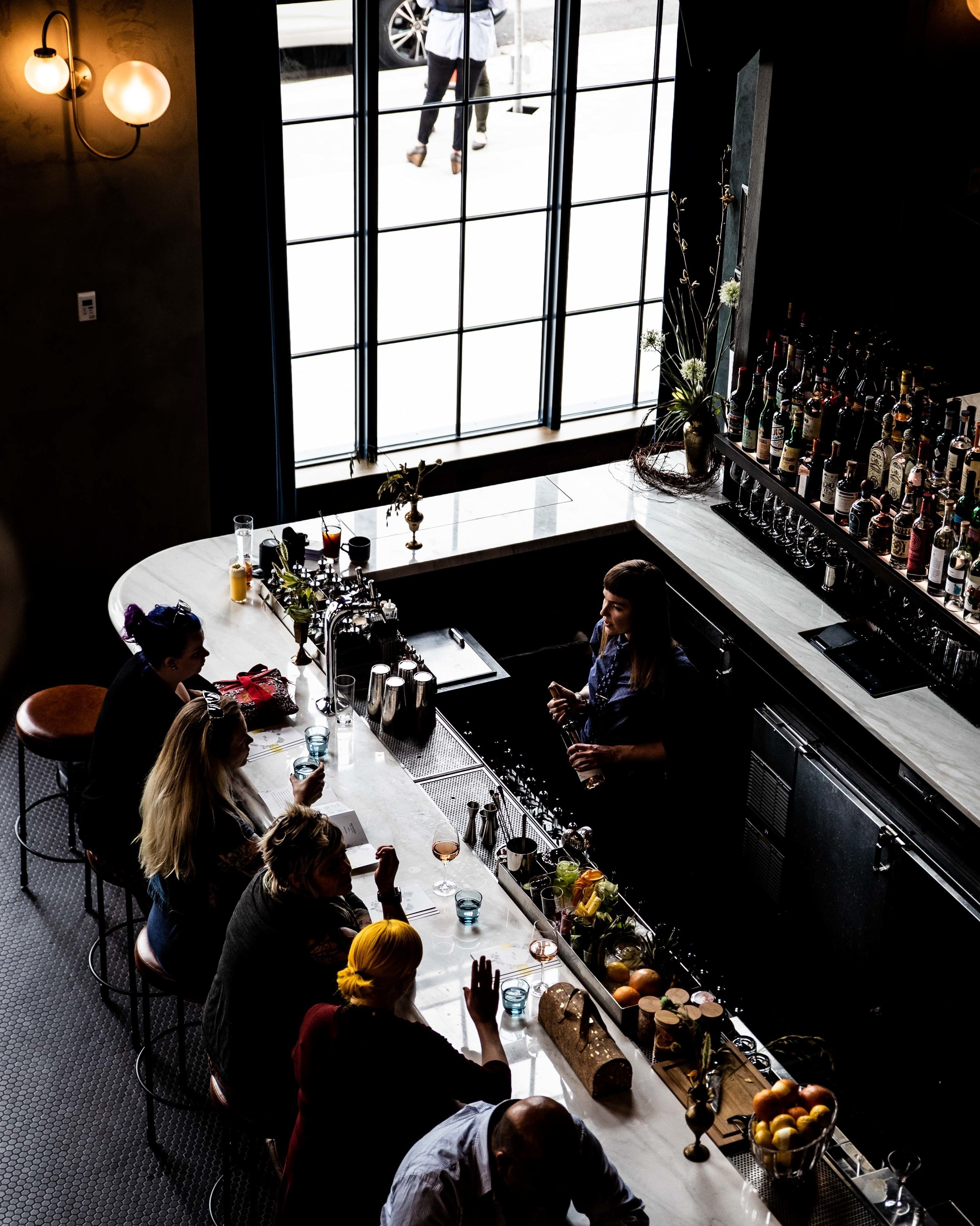 Eine Bar mit einer langen Theke, verschiedenen Flaschen und Gläsern. Personen sitzen an der Bar, und eine Person steht hinter der Theke.