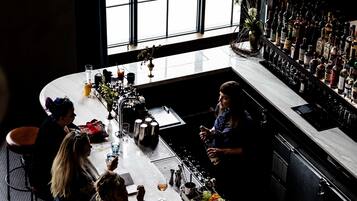 Eine Bar mit einer langen Theke, verschiedenen Flaschen und Gläsern. Personen sitzen an der Bar, und eine Person steht hinter der Theke.