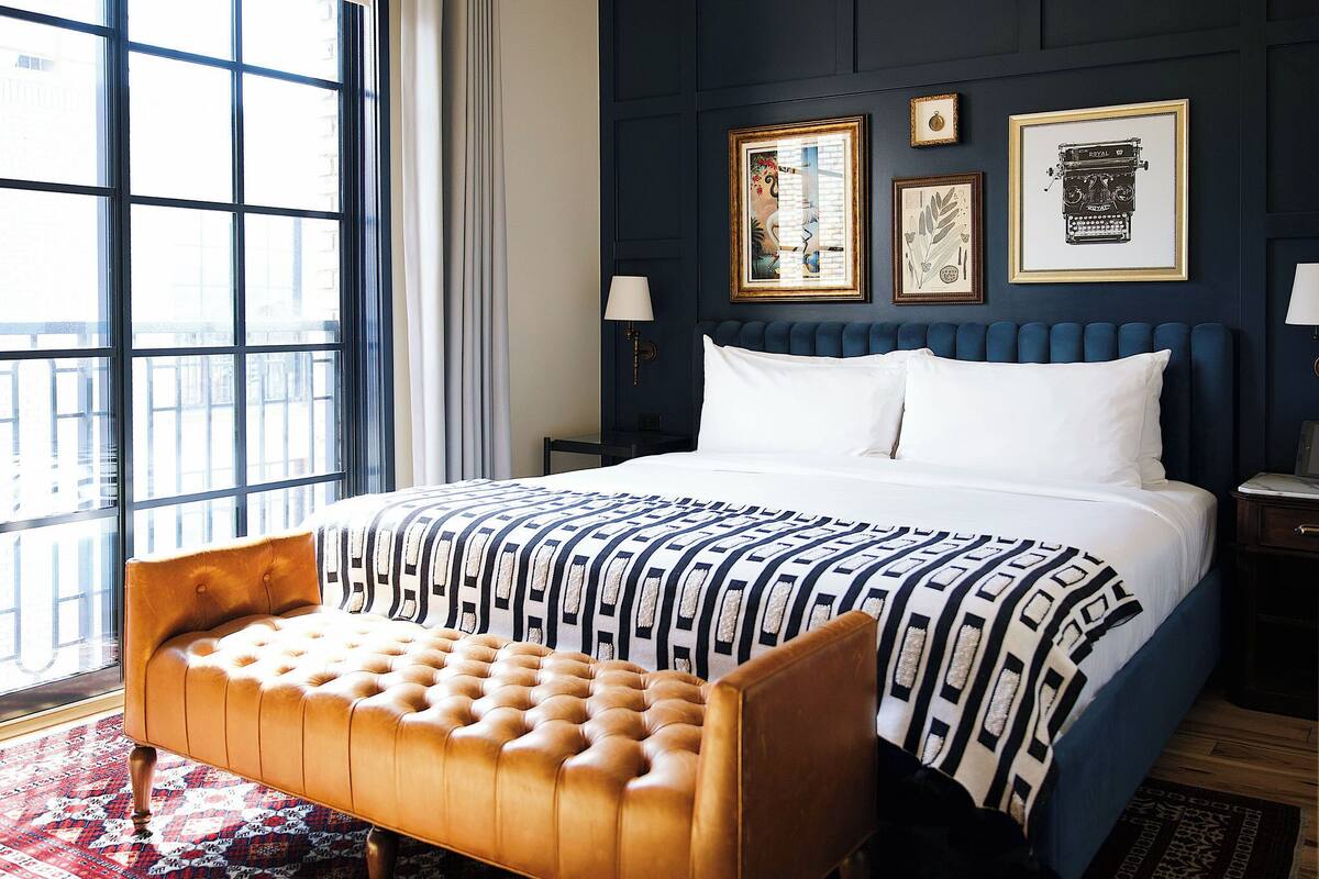 A bedroom with a large bed, a tufted bench, a dark wall with framed pictures, and a large window.